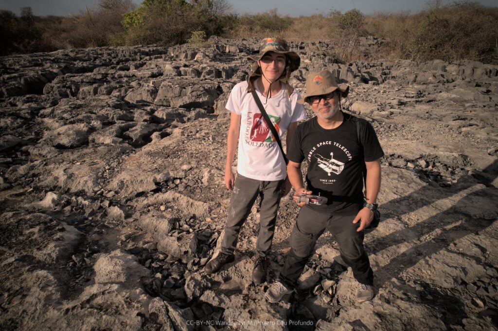 Wandeclayt do Projeto Céu Profundo, de barba e óculos, vestindo camiseta preta e chapéu. Henrique, guia do Lajedo, vestindo camiseta branca. Os dois homens olham na direção da câmera e se encontram sobre as rochas do Lajedo.