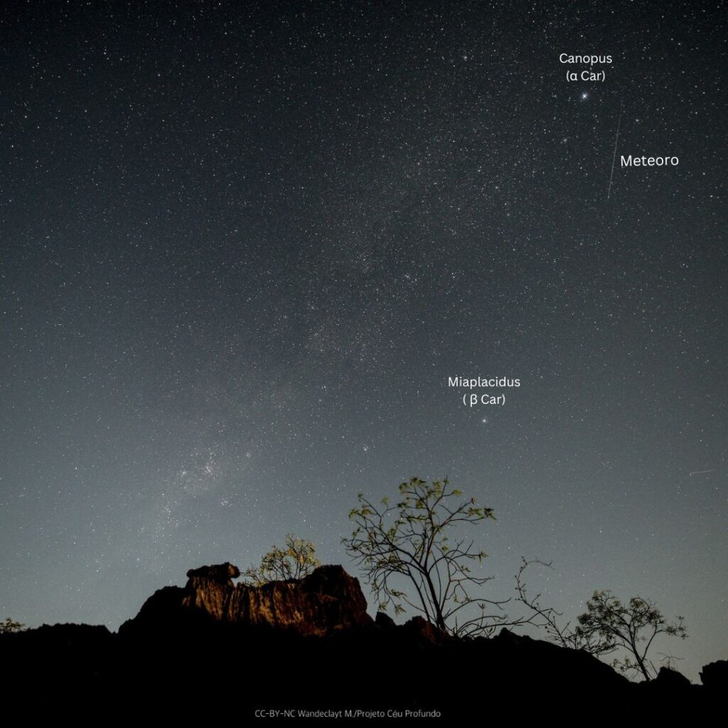 Um céu estrelado, com uma faixxa clara cortando a imagem diagonalmente. A faixa é a nossa galáxia, a Via Láctea. Um pequeno traço aparece num canto superior, formado pela passagem de um meteoro. Na parte baixo, uma formação rochosa iluminada pela Lua e algumas árvores delgadas.