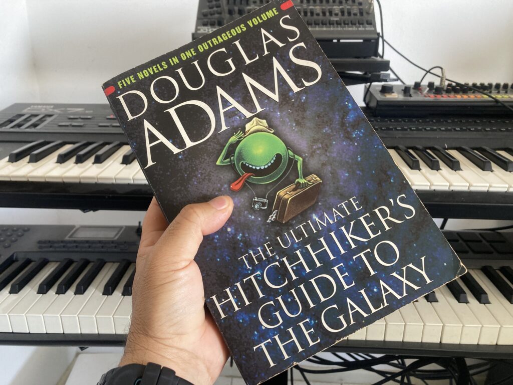 Pessoa segura um exemplar do livro The Ultimate Hitchhiker’s Guide to the Galaxy, de Douglas Adams. Ao fundo vê-se dois teclados e dois sintetizadores.