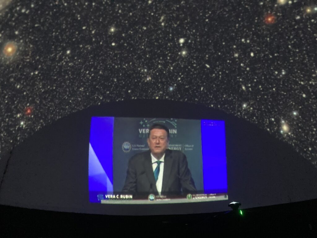Projeção na cúpula do planetário o evento que divulgou as primeiras imagens do Observatório Vera C. Rubin. No centro da imagem, aparece a projeção de um homem de terno falando ao microfone, com o fundo do palco exibindo logotipos e o nome do observatório. Ao redor, no teto da cúpula, vê-se a projeção de uma imagem astronômica com centenas de estrelas e galáxias sobre um fundo escuro. A sessão aconteceu no Planetário do Museu Interativo de Ciências (MIC), em São José dos Campos.