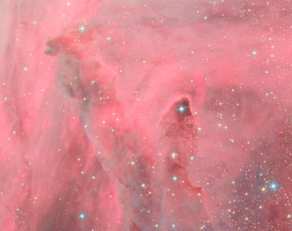 Detalhe da Nebulosa da Lagoa capturada pelo Observatório Vera C. Rubin. Na imagem, a nebulosidade é representada com uma coloração rosada e ao centro há duas regiões mais escuras trazendo um formato de pilar. Em primeiro plano, a imagem é composta de diversas estrelas com cores azuladas e alaranjadas.