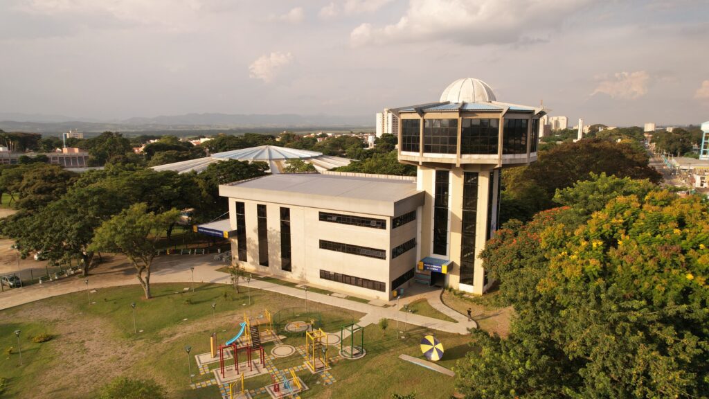 Vista aérea do Museu Interativo de Ciências. O edifício principal tem formato retangular com três andares e fachada clara. À direita, conectado ao prédio, há uma torre alta com uma cúpula no topo, onde está localizado o planetário. O entorno é arborizado, com árvores e gramados, e em frente ao prédio há um pequeno parquinho com brinquedos coloridos. Ao fundo, observa-se a cidade com edifícios e montanhas ao horizonte.