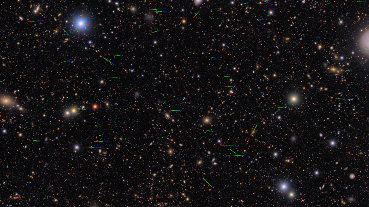 Imagem capturada pelo Observatório Vera C. Rubin, mostrando muitas estrelas e galáxias espalhadas pelo espaço de fundo escuro. Espalhados pela imagem, há dezenas de pequenos rastros coloridos — em vermelho, verde e azul — que indicam o movimento de asteroides registrados durante a exposição da imagem.